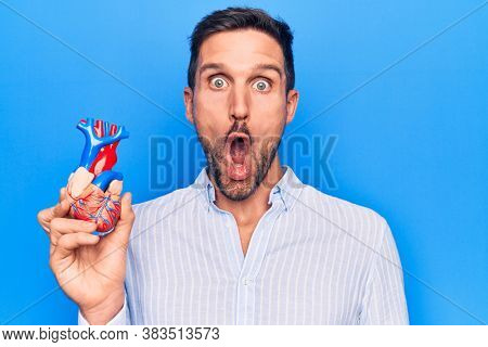 Young handsome man holding heart organ with veins and arteries over blule background scared and amazed with open mouth for surprise, disbelief face