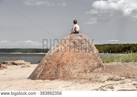 Boy Sunbathes On The Seashore, Sitting On A Large Granite Boulder