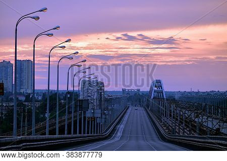 Sunrise Across The Bridge. View Of The Skyscrapers Of The City. The Column Of Street Lamps Is Illumi