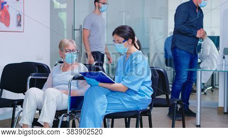 Disabled Senior Woman In Wheelchair Talking With Nurse That Wears Protection Mask Against Infection 