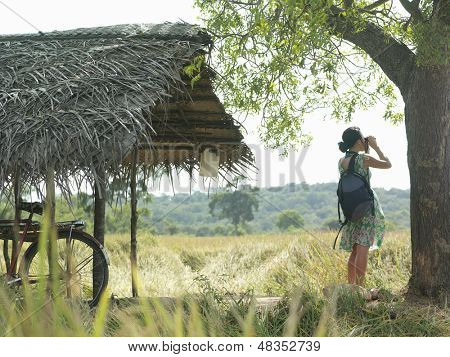 Side view of a young woman looking through binoculars with bicycle under thatched roof in field