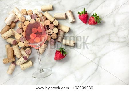 A high-key overhead photo of a glass of rose wine with corks and a fresh strawberries, on a white marble texture with a place for text