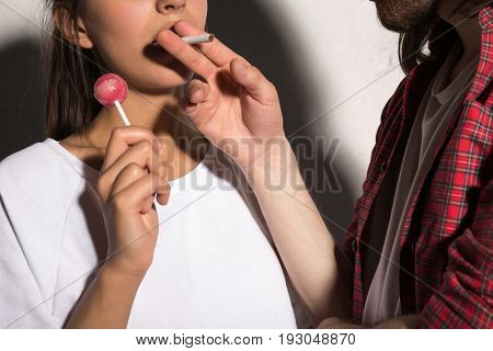 Cropped image of young hipster man standing over gray background give cigarette to girlfriend holding candy.
