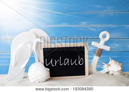 Chalkboard With German Text Urlaub Means Holiday. Blue Wooden Background. Sunny Summer Card With Holiday Greetings. Beach Vacation Symbolized By Sand, Flip Flops, Anchor And Shell.