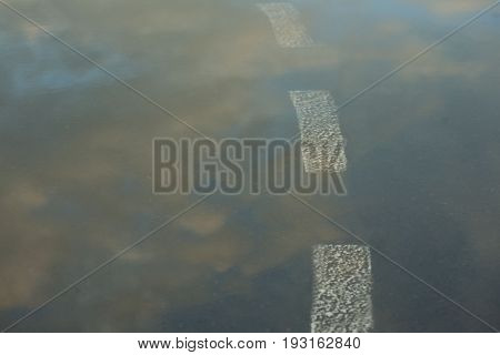 Reflection of the sky in the wet asphalt and puddle after the rain in the setting sun. Road marking going into the water. Close up view from the level of the dividing line. Road to the clouds