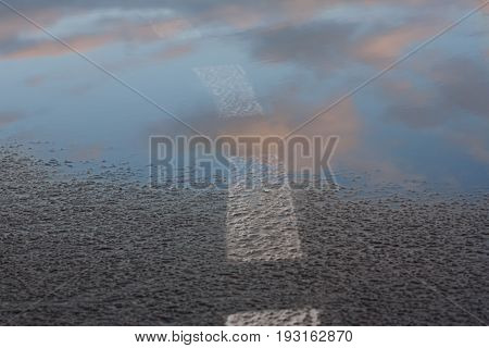 Reflection of the sky in the wet asphalt and puddle after the rain in the setting sun. Road marking going into the water. Close up view from the level of the dividing line. Road to the clouds
