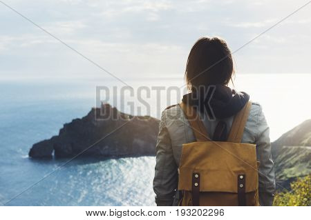 Hipster young girl with backpack enjoying sunset on seascape on peak mountain. Tourist traveler on background valley landscape view mockup. Hiker looking sunlight ocean in trip holiday in basque island Gaztelugatxe