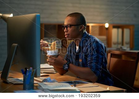 Profile view of handsome young man eating instant noodles with chopsticks while watching favorite TV series on computer, blurred background