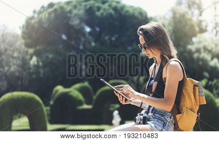 Hipster young girl with bright backpack and vintage camera using tablet or holding gadget planning travel plan. View tourist traveler on background Parc Laberint Barcelona. Mock up for text message
