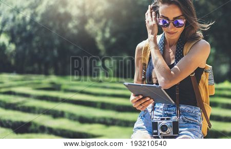 Hipster young girl with bright backpack and vintage camera using tablet or holding gadget planning travel plan. View tourist traveler on background Parc Laberint Barcelona. Mock up for text message