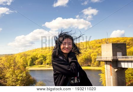 A Chinese woman with her hair blowing out of control on the lake side of Hop Brook Dam in Naugatuck connecticut on a sunny blue sky day.