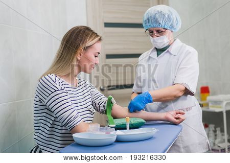 Preparation for blood test with pretty young blond woman by female doctor in white coat medical uniform on the table in white bright room. Nurse pierces the patient's arm vein with needle blank tube