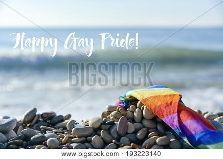 closeup of a rolled rainbow flag in a shingle beach, with the ocean in the background, and the text happy gay pride