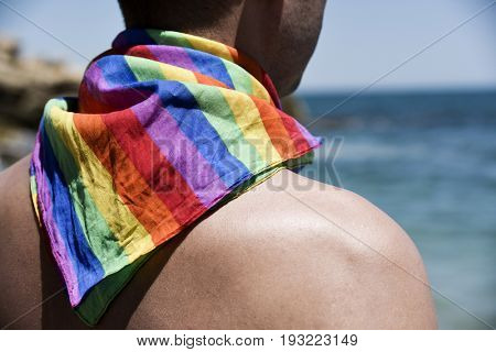a young caucasian man seen from behind with a rainbow patterned handkerchief tied around his neck in front of the ocean