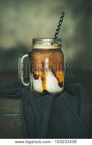 Iced caramel macciato coffee with milk in glass jar with straw on dark rustic wooden table, selective focus. Cold sweet refreshing summer drink concept