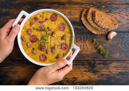 Smoked sausage and split pea soup with green thyme in a white pot in woman's hands the wooden rustic table top view.