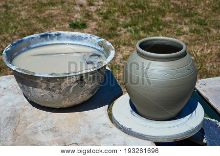 On the potter's wheel is a pot made of clay. On the table is a pelvis of water. In the water there are wooden template and a rubber scraper
