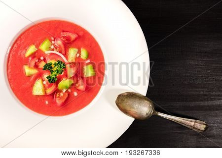 A closeup of a plate of gazpacho soup with slices of tomato, cucumber, and onion, with parsley leaves, on a dark background with a place for text