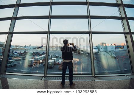Handsome man standing beside glass wall in modern airport terminal, taking photo picture of airplane aircraft, travelling to other countires