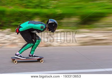 A young man in a fullface helmet and a leather suit in a special rack rides a longboard on afsaltu sunset in the background mountains and beautiful sky at high speed.