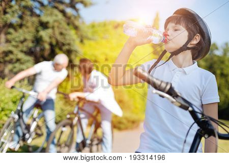 Nurturing moisture. Emotional engaged cool kid spending the day with his family and riding bikes with them while stopping for a gulp of water