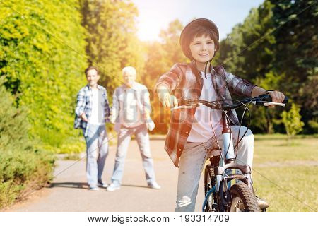 Important moment. Wonderful diligent active boy learning riding a bicycle while his family supporting him and giving some advice