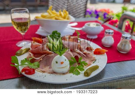 Stylish ham french fries and salad dish with funny decorated egg white wine glass mayonnaise salt and pepper on the background
