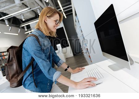   Smiling young lady with a leather backpack leaning on the display while testing the technological capabilities of a template personal computer.