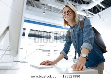  Low angle view of a smiling blonde woman leaning on a store display and looking at a personal computer while shopping at an electronics section of a local department store.