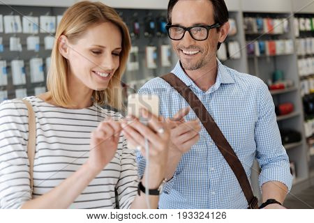  Excited couple grinning widely while testing all the electronics capabilities of the latest cellphone model while shopping at can electronics section of a local department store.