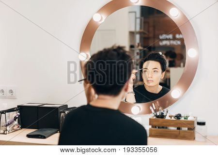Attractive woman primp at the mirror with lamps in the beauty studio. A happy satisfied customer of make-up and hairstyle service.