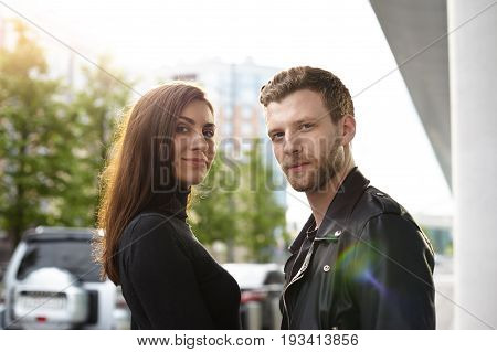 Waist up portrait of beautiful young Caucasian brunette female standing in urban environment close to her attractive stylish boyfriend both looking at camera. People style and fashion concept