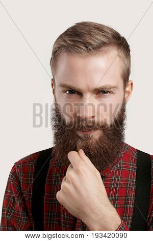 People and lifestyle. Fashion and style. Headshot of handsome young European macho man wearing treny clothing looking at camera deep in thoughts touching his fuzzy beard and squinting eyes
