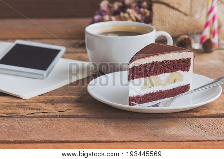 Banana chocolate cake on white plate served with black coffee. Chocolate cake layered with whipped cream and banana look so delicious. Enjoy during play smart phone with chocolate cake and coffee. Triangle slice piece of chocolate cake.