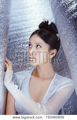 Creative art make-up and hairstyle. Beautiful young multi-racial asian caucasian woman. Beautiful young model Japanese style girl silver urban clothes with conceptual hairstyle and makeup against metallik silver wall looking at wall.