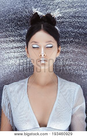 Creative art make-up and hairstyle. Portrait of asian young woman. Beautiful young model Japanese style girl silver urban clothes with conceptual hairstyle and makeup against metallik silver wall with closed eyes.
