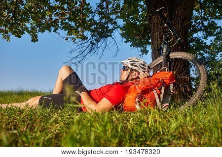 Traveler cyclist have a rest under oak tree with his bike behind him on summer afternoon. Mountain bikyclist in white helm and red t-shirt on green grass aganist blue sky. Travel concept.