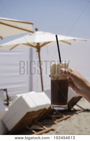 Iced coffee in hand with blurred beach background.
Female hand holding an iced glass of cappuccino freddo coffee with a black straw, on a sunny summer day.