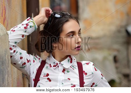 Closeup fashion portrait of young pretty hipster girl with dark brown hairs wearing stylish denim braces for trousers glasses and shirt with funny print cherry.Outdoors bakground.Copy space.Warm photo