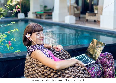 Sexy woman using laptop computer on a lounge near swimming pool outdoors. Tropical garden of Bali island.