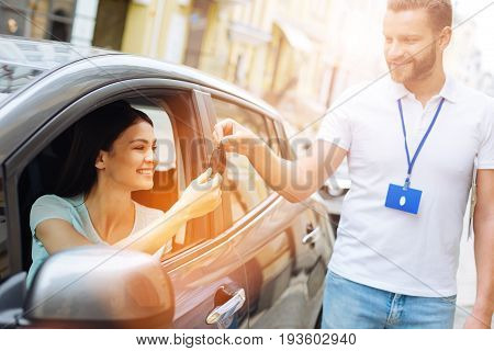 Amazing service. Handsome car rental agency employee handing car keys to a happy beautiful woman sitting in the driver seat and accepting them willingly