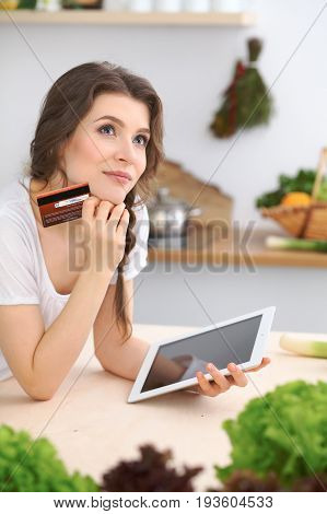 Young  woman looking for a new recipe for  cooking in a kitchen. Housewife is making online shopping by tablet computer and credit card.