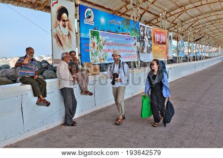 Hormuz Island Hormozgan Province Iran - 17 april 2017: Tourists go to the pier past banners with political and commercial advertising and three local old people.