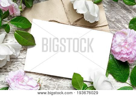 blank white greeting card and envelope in frame of roses flowers over white wooden background. mock up. top view. wedding invitation