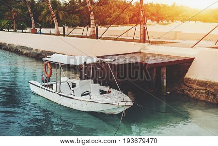 Small old used speedboat with awning parked in teal transparent water to wooden dock near quay wall with stairs with lanterns and palms sand beach with multiple chaise longues in background Maldives