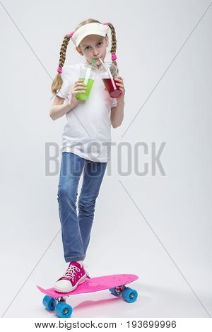 Kids Concepts. Portrait of Little Caucasian Blond Girl in Visor Posing on Pennyboard With Two Cups of Juice and Straw. Against White. Vertical Shot