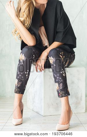 Young fashionable blonde woman in a black jacket in classic high-heeled pants sits on an ottoman