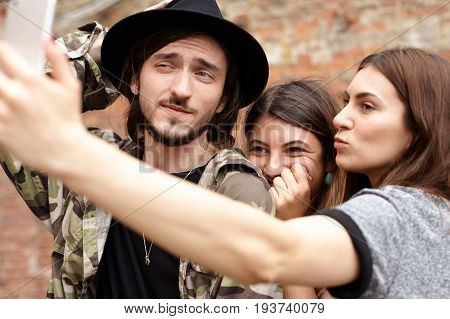 Happy fashionable young tourists dressed in stylish clothing posing for selfie looking at camera of smart phone smiling and making faces while sightseeing in foreign country during road trip