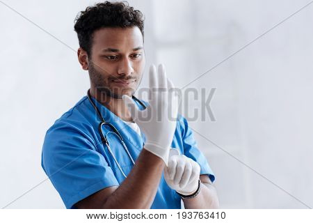 Visit doctor. Positive delighted man keeping smile on his face while wearing sterile gloves and standing over white background