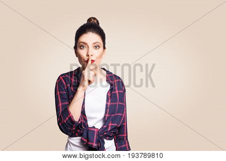 Portrait of beautiful young caucasian woman with black hair bun holding index finger at lips asking to keep silence or not tell anyone her secret raising brows saying 